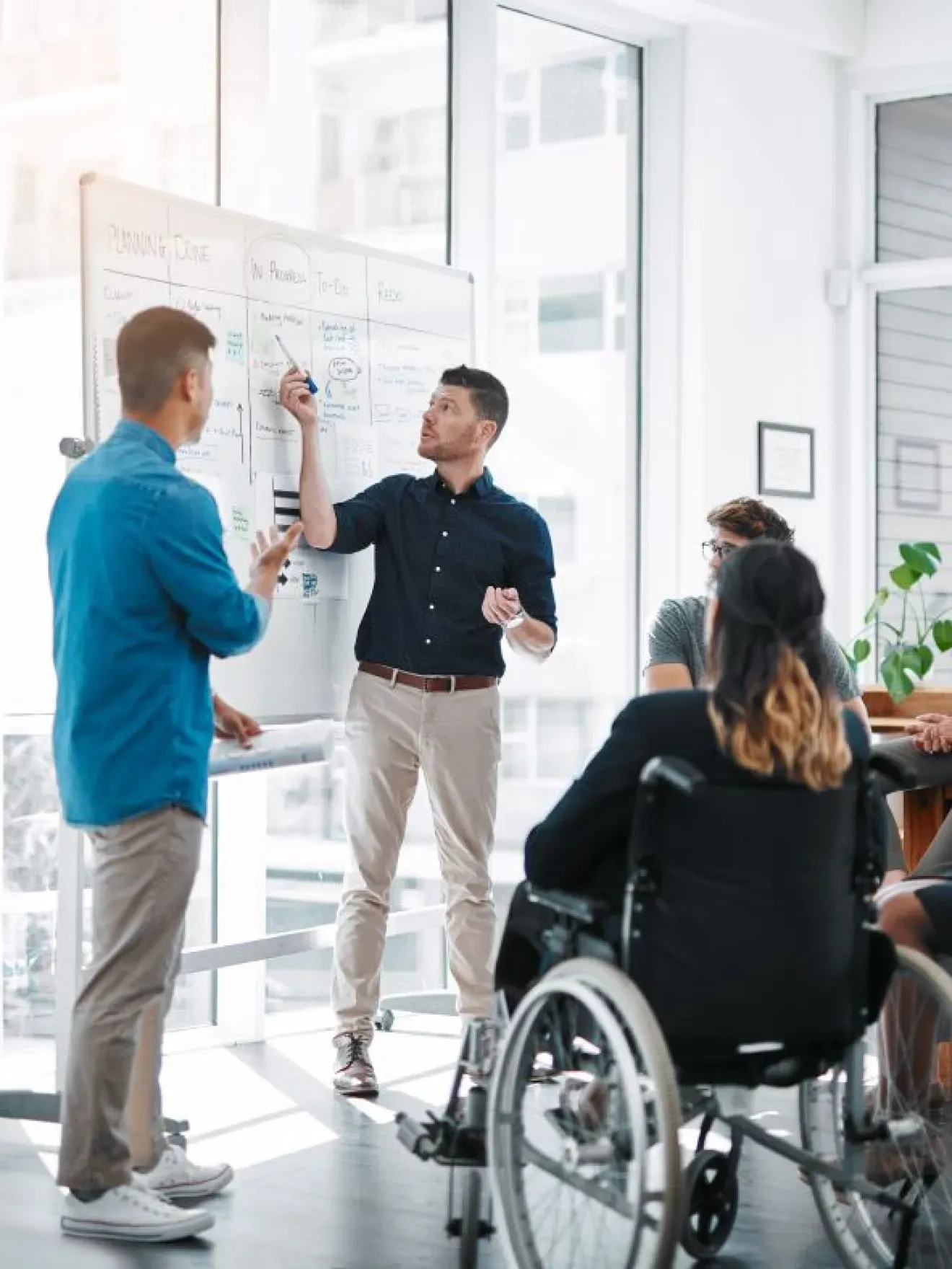 2 personnes en fauteuil roulant avec collègues regardent un tableau dans un bureau