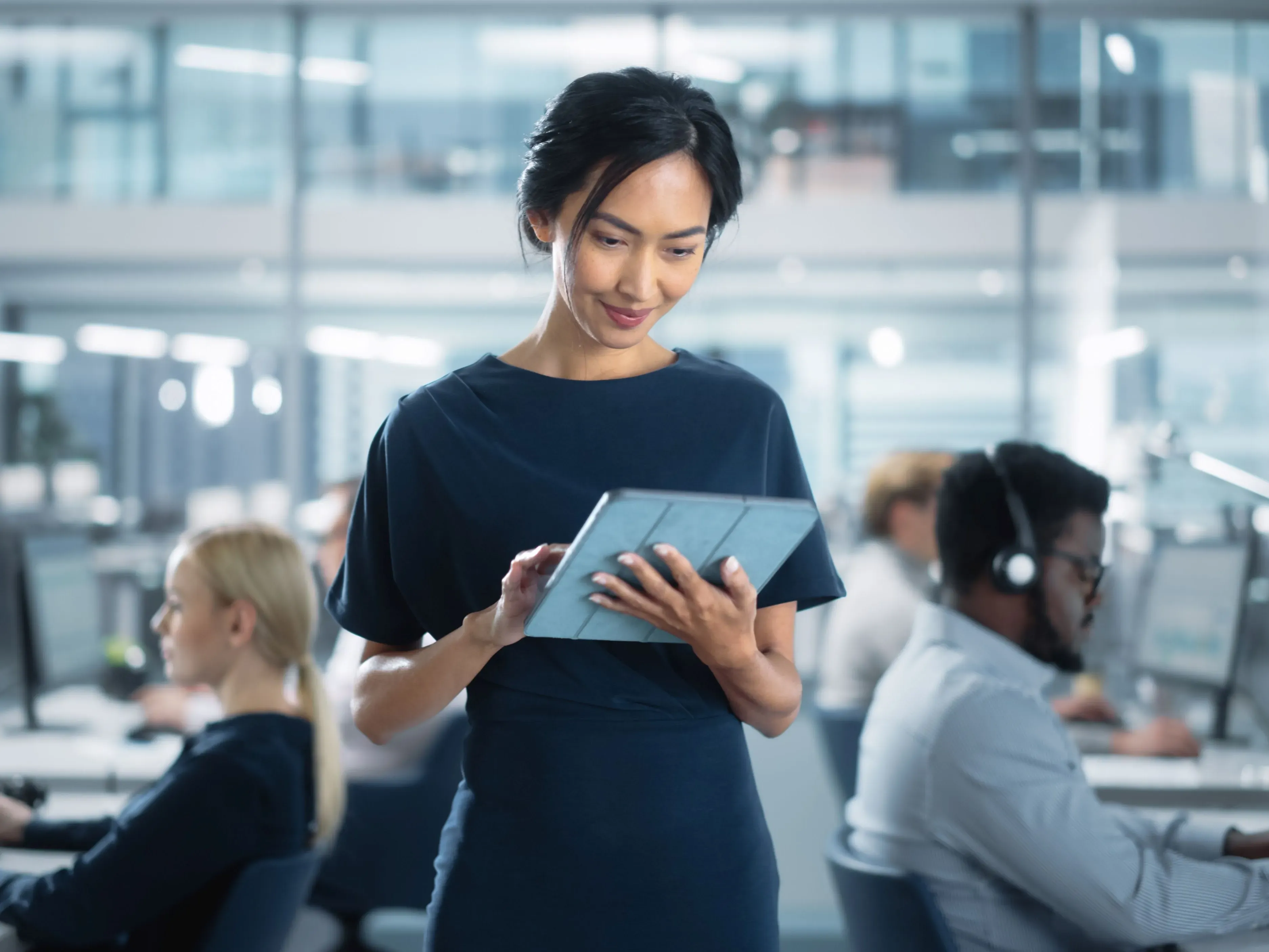 Jeune femme devant sa tablette dans un bureau où deux personnes travaillent dans le fond